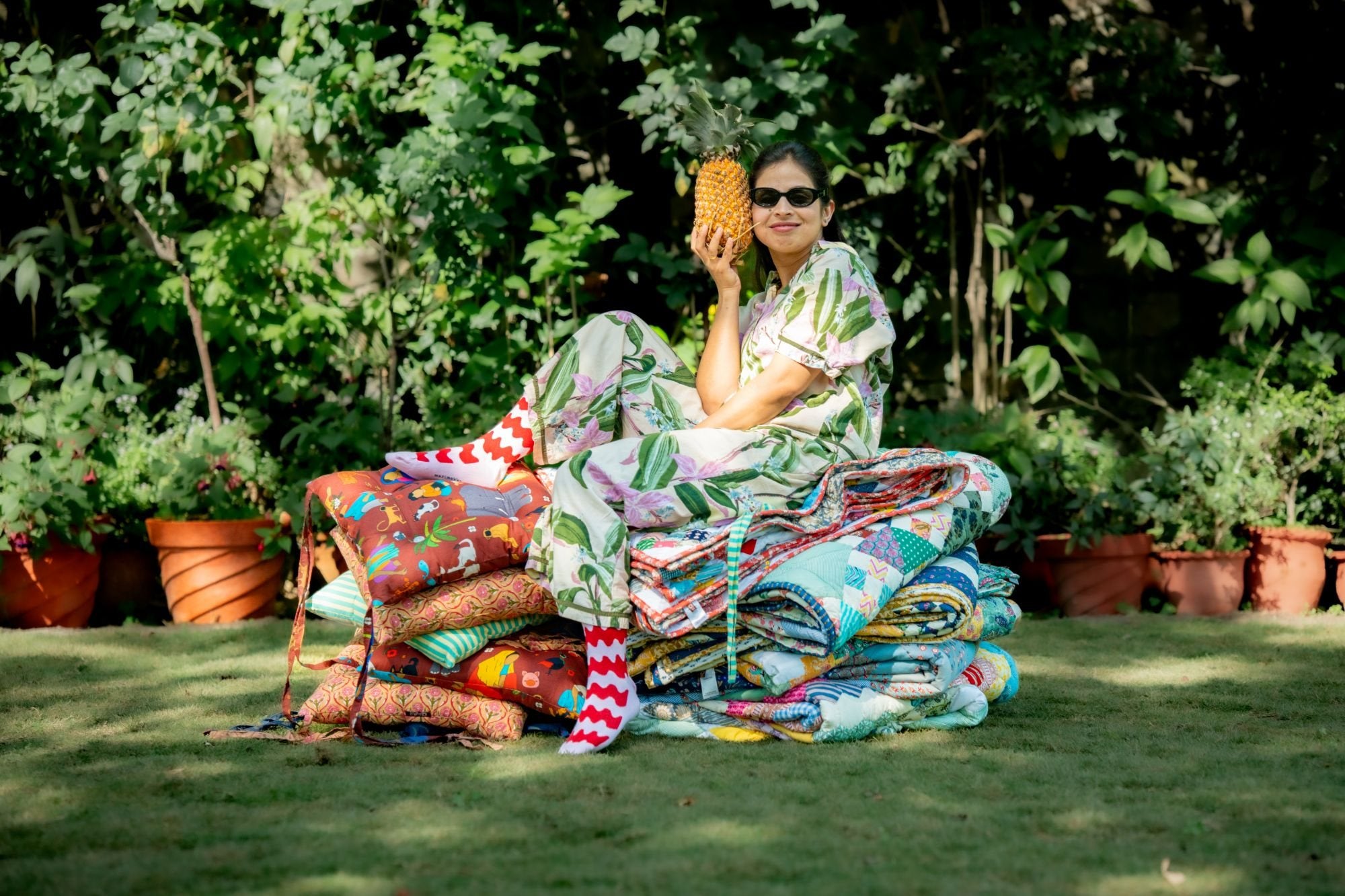 A woman in sunglasses holding a pineapple, sitting on a pile of patchwork quilts and printed cushions in a garden