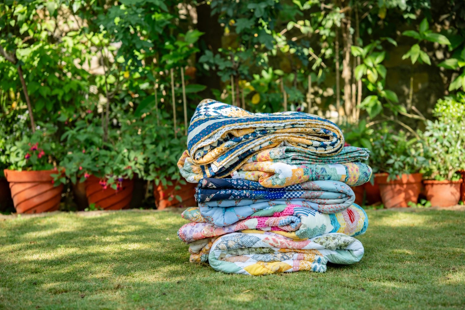A tall stack of folded patchwork quilts on a garden lawn, with potted plants behind