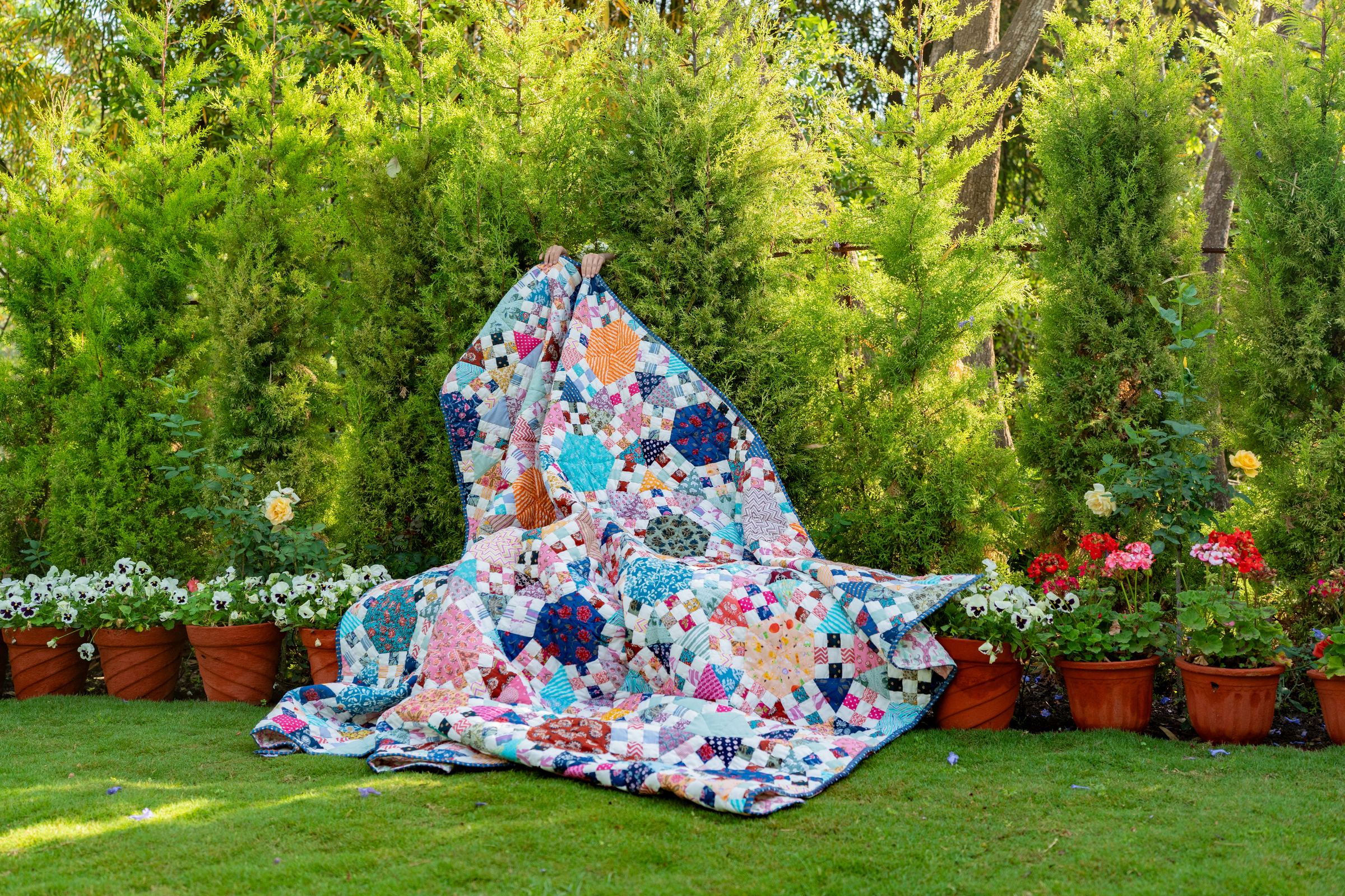 A patchwork quilt of hexagon flowers held up in a garden, among potted flowers